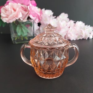 May include: A vintage pink glass sugar bowl with a lid. The bowl has two handles and a diamond pattern. The lid is topped with a small, pointed finial. The background features pink and white flowers in a glass vase.