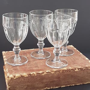 May include: Four clear wine glasses with a ribbed design on the bowl and stem. The glasses are arranged on a vintage book with a worn, brown cover. The background is a dark colour.