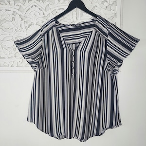 Puede incluir: Una blusa a rayas negras, blancas y grises con cuello en V y mangas cortas. La blusa tiene una botonadura frontal y está colgada de una percha negra. La tela tiene rayas verticales de diferentes anchos.