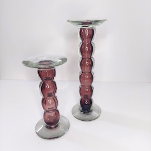 Peut inclure: Deux bougeoirs en verre. Les bases et les sphères empilées sont de couleur violet foncé, tandis que les dessus sont transparents. Le bougeoir le plus grand mesure environ 30 cm de haut, et le plus petit mesure environ 20 cm de haut. Les deux ont une base circulaire.
