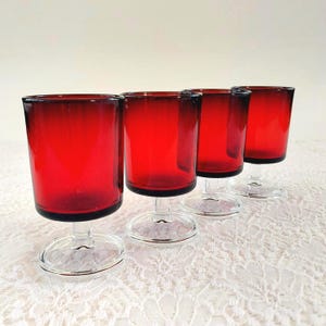 May include: Four vintage red glass cordial glasses. Each glass has a cylindrical bowl and a clear stem and base. The red glass has a dark rim. The glasses are arranged in a row on a white lace tablecloth.