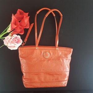 May include: A bright orange leather tote bag with a gold logo and a slightly textured surface.