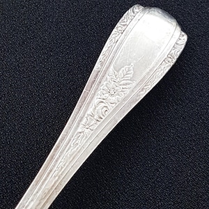 May include: Close-up of the ornate handle of a silver spoon. The handle is decorated with a floral design.