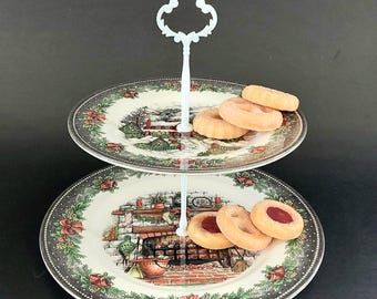 May include: A two-tiered porcelain serving stand featuring a white central handle and two round plates decorated with festive holiday scenes, including a snowy landscape and a fireplace, surrounded by green pine and red bow borders with cookies.