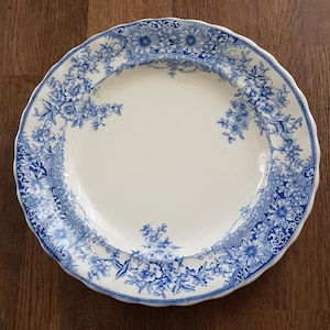 May include: A white ceramic plate with a blue floral design. The plate features a scalloped edge and a detailed pattern of flowers and leaves. The blue and white color scheme gives the plate a classic, vintage look. Ideal for serving food or as a decorative piece.