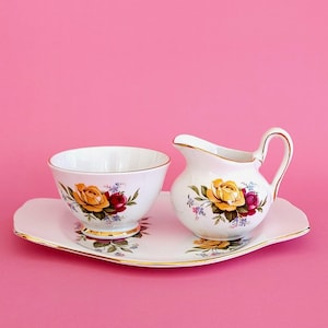 Peut inclure: Un ensemble sucrier et pot à lait en porcelaine blanche avec bordure dorée, orné de motifs floraux de roses jaunes et rouges. L'ensemble comprend un plateau en porcelaine blanche assorti aux bords dorés, décoré de petites fleurs bleues.