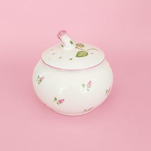 May include: A white ceramic potpourri jar with a rounded body and a matching lid. The surface is decorated with small pink rosebuds and green leaves. The lid features a sculpted pink rosebud handle and several small circular ventilation holes.