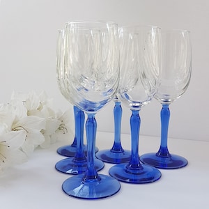 May include: Five clear glass wine glasses with blue stems. The glasses are arranged in a semi-circle on a white surface.