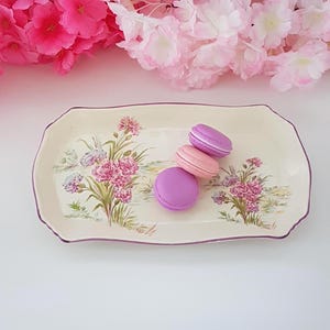 May include: A rectangular, cream-coloured ceramic serving dish with a floral design and a purple border. Three macarons in shades of pink and purple rest on the dish. Pink flowers are in the background.