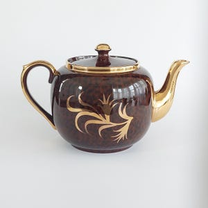 Peut inclure: Une théière ronde en céramique avec un motif marbré brun foncé et une finition brillante. La théière comprend une anse dorée, un bec verseur doré, un couvercle bordé d'or avec un bouton, et un motif floral doré sur le devant.
