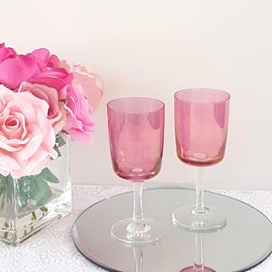 May include: Two pink wine glasses with clear stems and bases sit on a mirrored surface. A bouquet of pink roses in a clear square vase is to the left. The glasses reflect the light, creating a soft, elegant aesthetic.