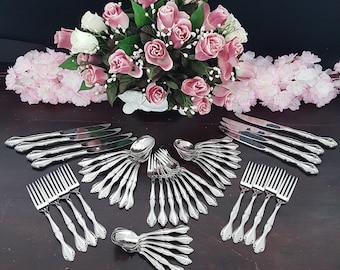 Könnte beinhalten: Eine Sammlung von silberfarbenem Besteck, darunter Gabeln, Messer und Löffel, angeordnet auf einer dunklen Holzoberfläche. Ein Strauß aus rosa und weißen Rosen und rosa Blumen befindet sich im Hintergrund.
