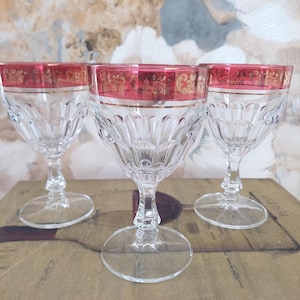 Può includere: Tre calici di cristallo trasparente con bordo rosso e design a filigrana dorata. I calici presentano un motivo dettagliato e strutturato sulla coppa e un gambo e una base trasparenti. Sono esposti su una superficie di legno.