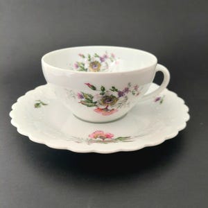 May include: A white porcelain teacup and saucer set featuring a delicate floral pattern with pink, purple, and green blossoms. The teacup has a curved handle, and the saucer has a scalloped edge with a subtle embossed dot texture along the rim.