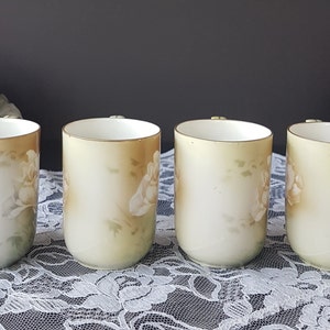 May include: Four white porcelain mugs with a floral design. The mugs have a light green and brown background with white flowers. The mugs are all the same size and shape.