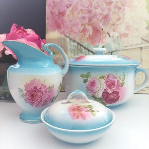 May include: A vintage porcelain tea set comprising a jug, a lidded pot, and a small bowl. The set is painted with a light blue and white gradient, accented with gold trim and pink rose floral designs. The set is ideal for serving tea.