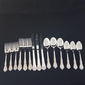 Pode incluir: Um conjunto de talheres prateados, incluindo garfos, facas e colheres, dispostos sobre uma superfície preta. Os talheres têm um padrão floral decorativo nas alças. O conjunto inclui garfos, facas e colheres de vários tamanhos.