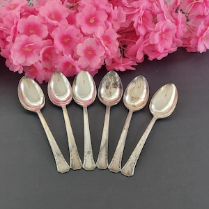 Könnte beinhalten: Ein Set aus sechs silbernen Löffeln, angeordnet auf einer dunklen Oberfläche, mit rosa Blüten im Hintergrund. Die Löffel haben ein klassisches Design mit detaillierten Griffen und ovalen Schalen. Ein Vintage-Besteckset.