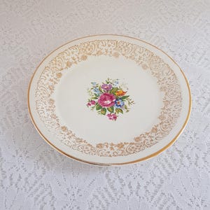 May include: A cream-coloured vintage plate with a gold rim and an ornate gold filigree border. The centre features a floral bouquet with pink, blue, yellow, and purple flowers. This decorative plate is ideal for serving or display.