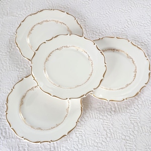 May include: Four white ceramic plates with scalloped edges and gold trim. Each plate features a delicate gold filigree design around the inner rim. The plates are arranged on a white textured surface.