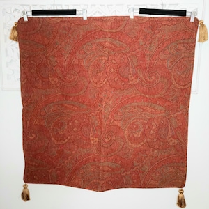 May include: A rectangular, red paisley patterned textile with gold tassels at each corner. The fabric is hung from a black bar with white clips. The paisley design features swirls and floral motifs in shades of red and brown.