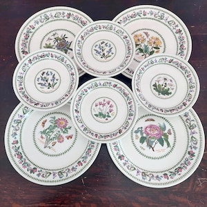 Könnte beinhalten: Eine Sammlung weißer Keramikteller mit botanischen Illustrationen. Jeder Teller zeigt ein zentrales Blumendesign und einen Rand aus grünen Blättern und kleinen Blüten. Die Teller variieren in der Größe und zeigen verschiedene Blumen- und Schmetterlingsmotive.