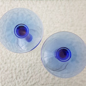 May include: Two blue glass goblets with a clear base and a blue rim. The goblets are on a white textured surface.