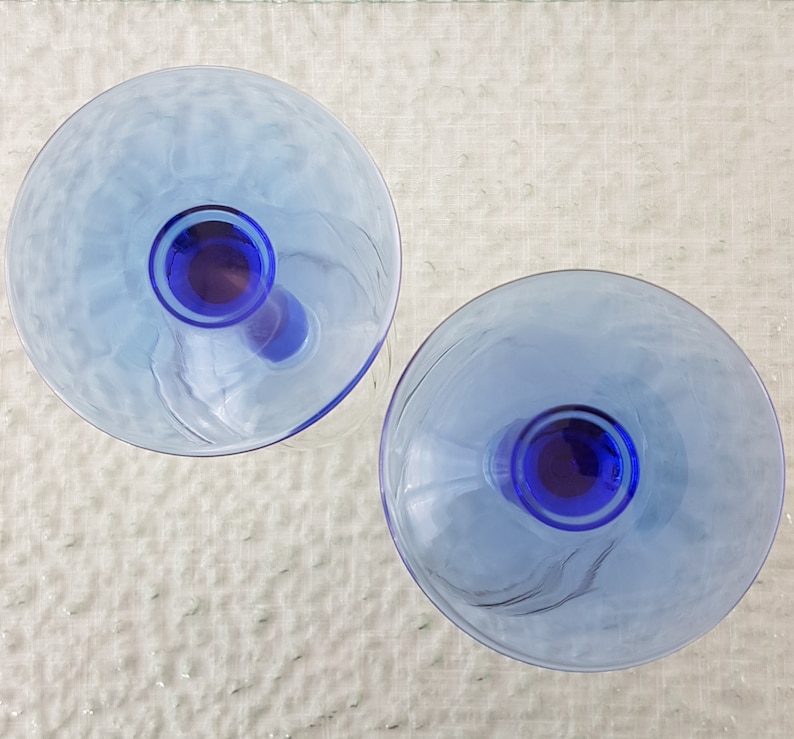 May include: Two blue glass goblets with a clear base and a blue rim. The goblets are on a white textured surface.