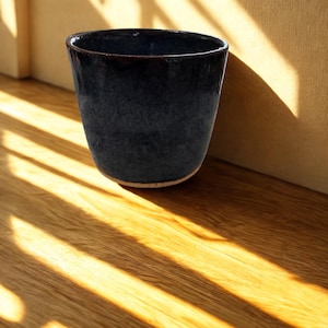 May include: A dark blue ceramic cup with a textured surface. The cup is cylindrical with a slightly flared rim and sits on a light-colored base. The cup is positioned on a wooden surface, with sunlight and shadows from a window creating a pattern.