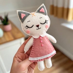 May include: A handmade felt cat doll. It has a white head, grey ears with pink accents, and a pink dress. The doll has closed eyes, pink cheeks, and an orange cross-stitch detail. The dress has a white collar and pink buttons.