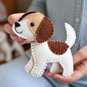 Puede incluir: Un pequeño adorno de perro hecho a mano en fieltro con manchas blancas y marrones. El peluche tiene detalles de bordes cosidos, un ojo de cuenta negra y un pequeño collar rojo. El perro aparece de pie con la cola levantada y orejas caídas.