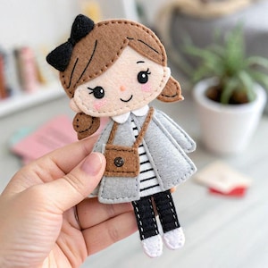 Puede incluir: Muñeca de fieltro hecha a mano con cabello castaño, un lazo negro y un abrigo gris. La muñeca tiene un pequeño bolso marrón y lleva una camisa a rayas, pantalones negros y zapatos blancos. La cara de la muñeca tiene mejillas rosadas y una expresión sonriente.