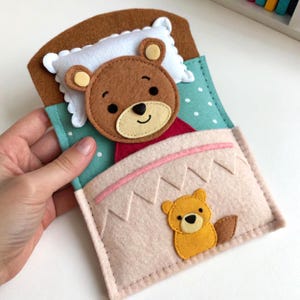 May include: A handmade felt toy depicting a bear in bed. The brown bear is nestled under a pink blanket with a smaller yellow bear at the foot of the bed. The bed is on a teal background with white polka dots, and a white pillow.