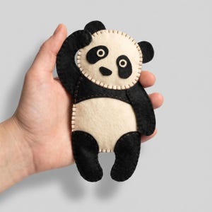 May include: A handmade panda bear plushie made of black and cream-colored felt. The panda has black eyes, ears, and limbs, with a cream-colored face and belly. The plushie is stitched together with a contrasting thread and has a hand-stitched appearance.