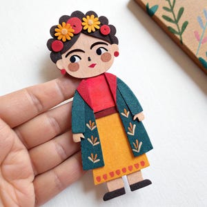 May include: A wooden brooch inspired by Frida Kahlo. The brooch shows a stylized Frida with a floral crown, red earrings, and a colourful outfit. The brooch is made of layered wood and felt.