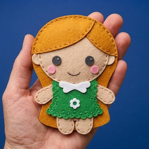 May include: A handmade felt doll with a tan face, orange hair, and a green dress. The doll has pink cheeks, black eyes, and a white bow. The doll is approximately 4 inches tall.