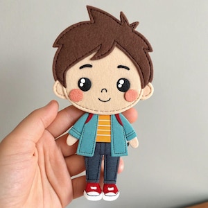 May include: A flat felt doll featuring a cartoon character with brown hair, large black eyes, and pink cheeks. The figure wears a teal jacket over a yellow and white striped shirt, dark gray pants, and red sneakers with white soles and laces.