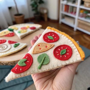 May include: Felt pizza slice with red tomato slices, green basil, and a tan cheese wedge. The crust is yellow and the base is off-white. Other felt pizza slices are in the background.