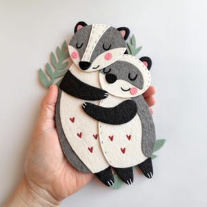 May include: A felt art piece featuring two hugging badgers. The badgers are primarily gray and white with black accents and pink cheeks. Small red hearts are scattered on the white bellies. Green leaf accents are visible.