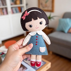 Peut inclure: Poupée en feutre faite à la main, avec une coupe au carré brun foncé et un nœud rose. La poupée porte une robe bleue avec un col blanc, des boutons rouges et des chaussures rouges. La poupée mesure environ 20 cm de haut.