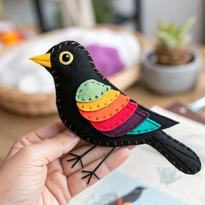 May include: A handmade felt bird with a black body, yellow beak, and colorful rainbow-colored wing accents. The bird has a stitched design and wire legs. The bird is held in a hand.