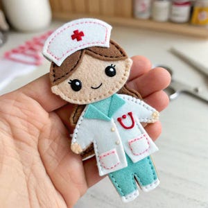 Puede incluir: Muñeca de enfermera de fieltro con uniforme blanco, pantalón turquesa y gorro blanco con cruz roja. La muñeca tiene cabello castaño, una cara sonriente y ojos negros grandes. El uniforme de la enfermera tiene un estetoscopio rojo.