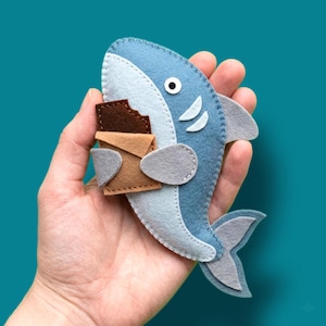 May include: A handmade felt shark in shades of blue, gray, and white, holding a brown chocolate bar. The shark has a white belly, gray fins, and a single black eye. The background is a solid teal color.