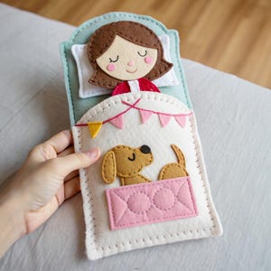 Op de afbeelding: Een handgemaakt vilten speelgoed dat een meisje voorstelt dat met een hond in bed slaapt. Het speelgoed heeft een lichtblauw hoofdbord, een wit kussen en een roze deken. De hond is bruin en het meisje heeft bruin haar.