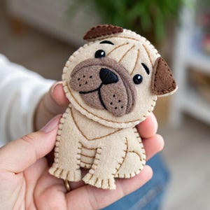 Könnte beinhalten: Ein handgefertigter Mops aus Filz in Beige und Braun. Der Mops hat ein detailliertes Gesicht mit schwarzen Augen und einer braunen Nase. Der Filzhund ist mit braunem Faden genäht, was ihm ein charmantes, handgefertigtes Aussehen verleiht.