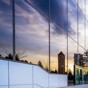 Op de afbeelding: Een reflectie van een klokkentoren met een witte wijzerplaat en zwarte wijzers is zichtbaar in een groot glazen gebouw. Het gebouw heeft een witte basis en een grijze betonnen vloer.