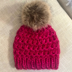 Puede incluir: Un gorro de punto de color rosa brillante con un gran pompón de piel sintética marrón en la parte superior.