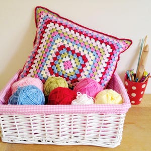 May include: A white wicker basket with a pink gingham lining holds colorful yarn balls and a crocheted pillow. The pillow features a square pattern in various colors, with a red border. A red polka-dot cup with crochet hooks is also visible.