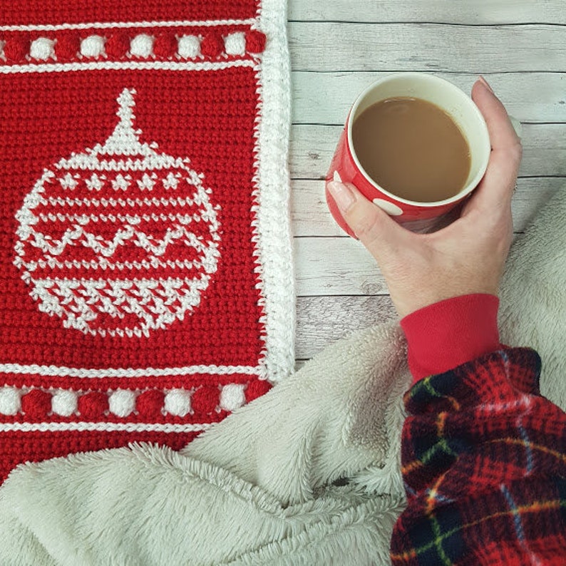 May include: A red and white crocheted blanket with a white border and a red and white Christmas ornament design. A hand is holding a red mug with a white rim and a brown liquid inside. The mug is sitting on a white fuzzy blanket.