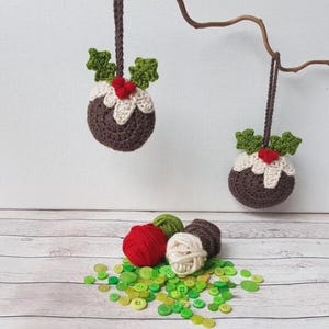 May include: Two crocheted Christmas pudding ornaments hanging from a branch with green holly leaves and red berries.  There are also balls of yarn and green buttons on a white wooden surface.
